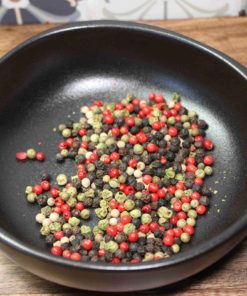 Assortiment de grains de poivre rouge, noir, jaune et vert dans un bol noir sur une table en bois.