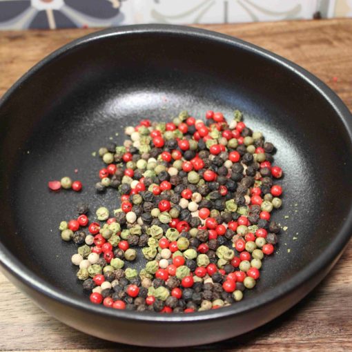 Assortiment de grains de poivre rouge, noir, jaune et vert dans un bol noir sur une table en bois.