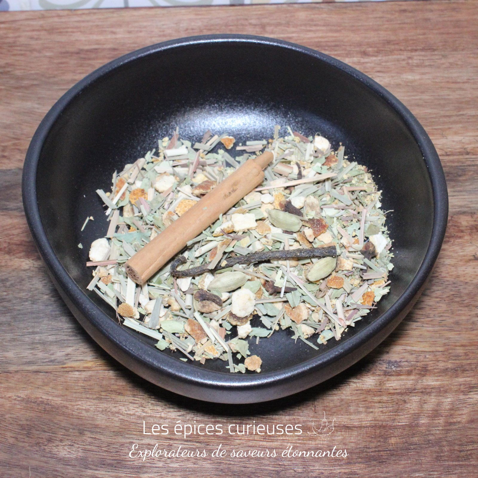 Préparation pour rhum Arrangé Marie Galante – Les épices curieuses Bol d'épices diverses sur une table en bois, incluant une branche de cannelle, cardamome et herbes séchées.