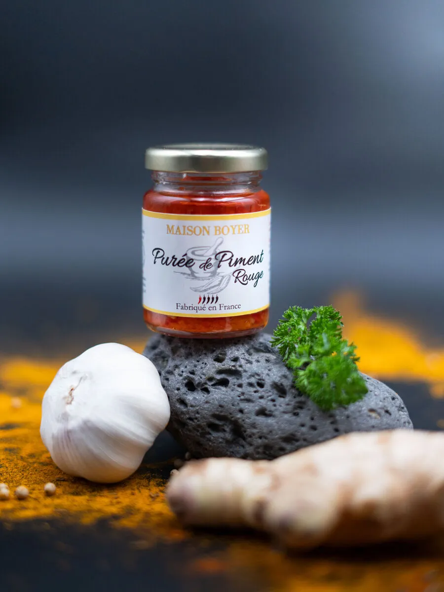 scene-puree-de-piment-maison-boyer-1.webp Bocal de purée de piment rouge Maison Boyer avec ail, gingembre et persil, fond coloré aux épices.